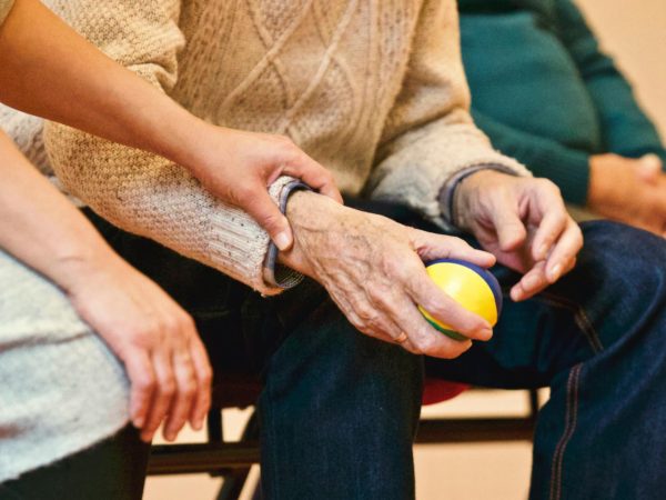 Una persona joven sujeta con cuidado la mano de una persona mayor que sostiene una pequeña pelota de colores en la otra mano, ambos sentados uno al lado del otro. La escena refleja cuidado y apoyo.